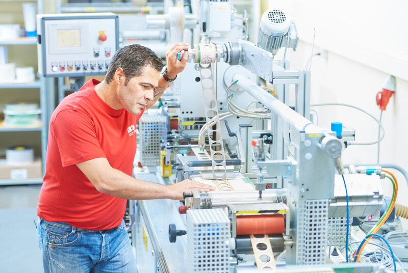 L'image montre la machine pour le découpage à plat, qui est utilisée par un homme portant un t-shirt rouge de l'IBZ. La bande estampée comporte des cercles.