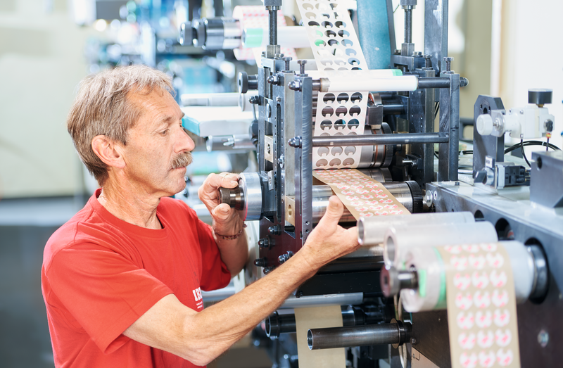 Sur l'image de la découpe rotative, on voit à gauche un employé en t-shirt rouge de l'ibz qui utilise la machine de découpe rotative. Sur la droite, on voit comment des cercles sont découpés dans la bande.