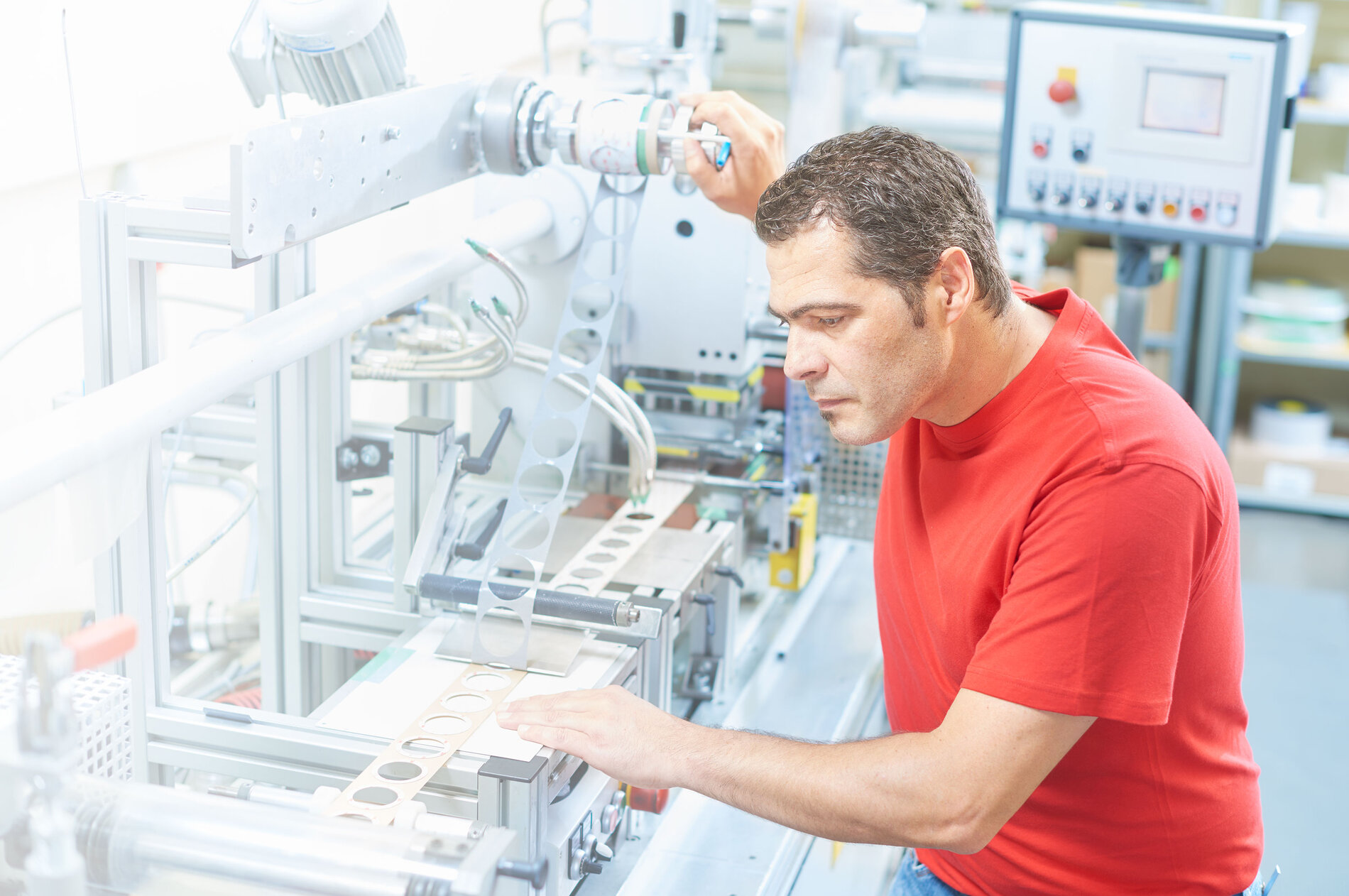 Sur l'image, on peut voir la production de pièces découpées par une machine utilisée par un ouvrier. Celui-ci est vêtu d'un t-shirt rouge IBZ. La machine imprime des pièces rondes